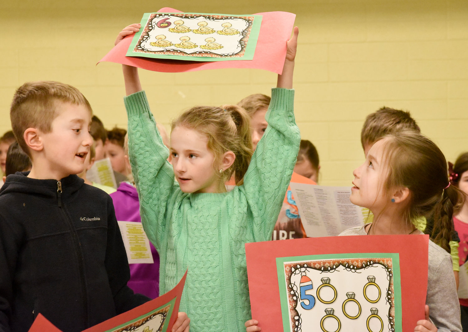 Kinsley Ginderich holds up a poster for six geese a laying providing musical accompaniment were Liam Frederick (left) and Sophia Patterson. The Mid-Prairie East second graders performed Christmas songs for the Rotary Club of Kalona on Dec. 17.